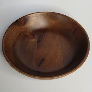 May include: A round, shallow wooden bowl with a smooth, polished finish. The bowl features a rich, dark brown colour with visible natural wood grain patterns and several dark, irregular knots embedded within the surface of the interior basin.