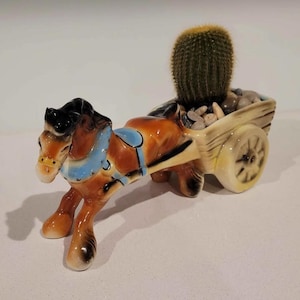 May include: A ceramic horse-drawn cart planter, with a brown and black horse and a blue harness. The cart carries a green cactus and small stones. The planter is cream and brown in colour.