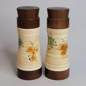 May include: A pair of cylindrical salt and pepper shakers featuring dark brown wooden caps and bases. The central bodies are cream-coloured with horizontal tan stripes and a painted pattern of green and orange leaves with black outlines.
