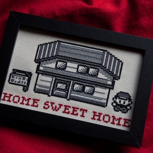 Puede incluir: Un bordado de punto de cruz enmarcado en negro, que representa una casa pixelada con un cartel que dice "HOME SWEET HOME" y un personaje pixelado de pie frente a ella.