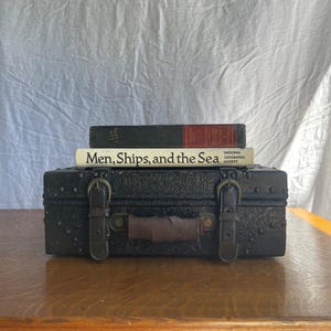 May include: A dark brown rectangular storage box with an embossed floral pattern, two front buckle straps, and a brown handle. Two books are stacked on top: a dark book with red spine details and a white book titled Men, Ships, and the Sea.