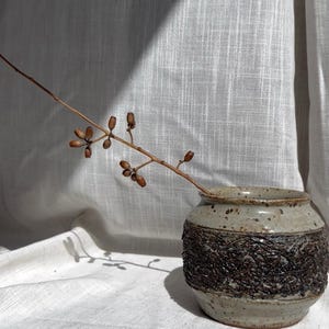 May include: A small, speckled ceramic vase with a textured, dark brown band around the middle. A slender branch with small, brown seed pods extends from the vase. The vase sits on a white linen surface.