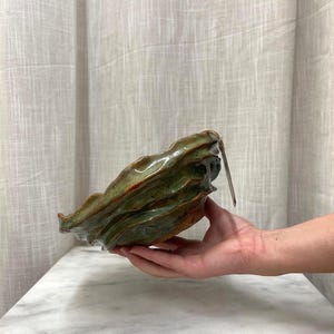 May include: A hand-formed ceramic bowl with an irregular, wavy rim and a glossy glaze featuring mottled shades of forest green, olive, and earthy brown. The bowl has a textured, organic surface and a small rectangular tag attached to the side.