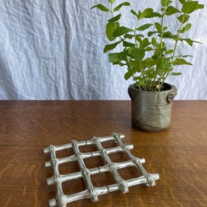 May include: A silver-toned metal trivet featuring a grid pattern designed to resemble bamboo stalks. The trivet is square-shaped with four horizontal and four vertical bars intersecting to form a lattice structure with raised nodes at each joint.