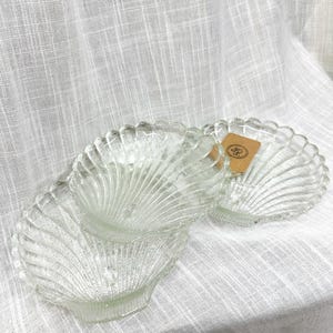 May include: Clear glass seashell-shaped dishes. The dishes have a scalloped edge and a textured, ribbed design. The image shows three dishes, with one partially stacked on another. The dishes are on a white, textured surface.