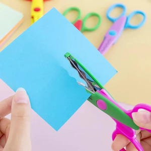 May include: A pair of pink and green decorative scissors cutting a blue square of paper. The scissors have a wavy blade design. Other colorful scissors are visible in the background.