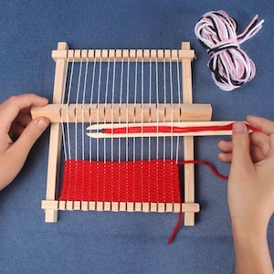 May include: A wooden weaving loom with white vertical threads and a red woven section. A wooden shuttle holds red yarn, and a ball of pink, black, and white yarn sits nearby. The loom is being used to create a small woven piece.