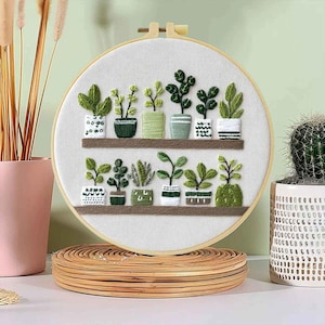 May include: Embroidered hoop art featuring a collection of potted plants on shelves. The plants are in various shades of green, with pots in white, green, and brown. The artwork is set against a white background within a wooden hoop, creating a botanical theme.
