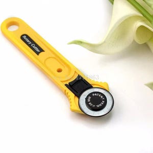 May include: A yellow rotary cutter with a black handle accent and a circular blade. The blade features the text 28 MM PATENT PEND, and the handle is labeled Rotary Cutter. The tool includes a circular hole at the top for hanging storage.