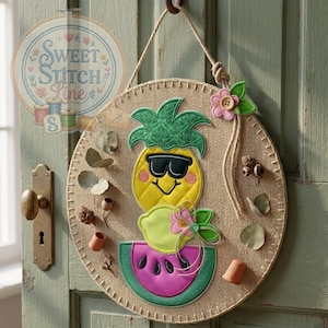 Op de afbeelding: Ronde wanddecoratie van jute met een lachende ananas met zonnebril, een citroen en een plak watermeloen. Het ontwerp bevat een groene ananas top, een roze bloem en de tekst "Sweet Stitch Line".