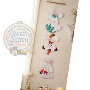 Pode incluir: Uma decoração de parede com três coelhos brancos com detalhes de cenoura. Cada coelho tem um laço de cor diferente e está suspenso de um pino de madeira. Um bastidor de ponto cruz com o texto "Sweet Stitch Line" também é visível.