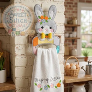 Pode incluir: Um porta-toalhas decorativo com tema de Páscoa com um coelho de feltro cinza com detalhes florais e uma gravata borboleta amarela. O suporte tem um design de patchwork colorido e contém uma toalha branca bordada com "Happy Easter" e detalhes florais.
