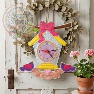 Pode incluir: Um relógio decorativo de feltro em forma de casa de pássaros com laço rosa, telhado amarelo e mostrador. Dois pássaros roxos e rosa ladeiam uma faixa com a palavra "Love". O relógio está pendurado numa porta branca.