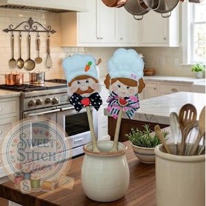 May include: Two wooden spoon decorations with chef designs. One has a moustache and bow tie, the other a watermelon scarf. Both have chef hats and are in a cream ceramic jar on a wooden surface.
