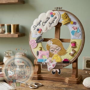 May include: A decorative embroidery hoop featuring a cartoon seamstress with glasses, a pink sewing machine, and the words "Design Create Inspire." The hoop includes fabric scraps, buttons, and a measuring tape design. It is displayed on a wooden stand.
