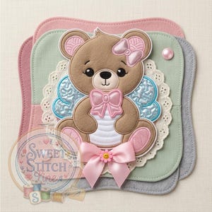 May include: A brown teddy bear applique with a pink bow on its head and chest, and blue wings. The bear is on a layered background of pink, green, and gray felt. A pink bow with a yellow smiley face is at the bottom. The image includes the text "Sweet Stitch Fine".