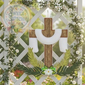 Könnte beinhalten: Ein Holzkreuz mit einem weißen, drapierten Tuch und grün-weißen floralen Akzenten. Das Kreuz hängt an einem weißen Gitterzaun, umgeben von weißen Rosen und Grün. Das Logo von Sweet Stitch Vine befindet sich in der oberen linken Ecke.