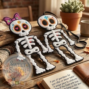 Pode incluir: Dois ornamentos de esqueleto do Dia dos Mortos. Um tem um laço rosa, o outro uma cruz. Ambos têm rostos de caveira detalhados com detalhes em laranja e preto. Os esqueletos são brancos com contornos pretos e detalhes de coração vermelhos.