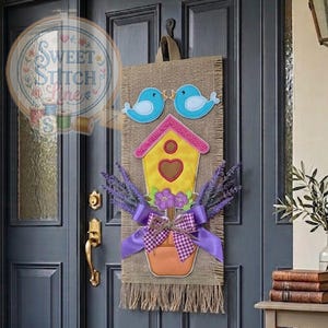 Può includere: Un'applique per porta in juta con una casetta per uccelli, due uccelli blu e un vaso di lavanda. La casetta è gialla con un tetto rosa e un ritaglio a forma di cuore. Un fiocco viola e frange completano il design.