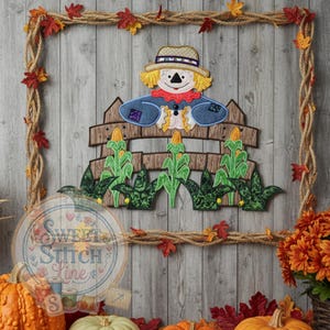 Pode incluir: Decoração de parede com tema de outono com um espantalho. O espantalho usa um chapéu de palha e roupas azuis, em pé atrás de uma cerca de madeira com plantas de milho. A moldura é feita de corda e decorada com folhas de outono.