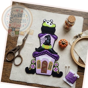 May include: An embroidered patch featuring a light purple house with a dark purple roof, a black witch hat, and a green cauldron on top. The house has a brown door, yellow windows, and black boots with purple and white candy accents at the base.