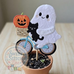 以下が含まれることがあります： ハロウィーンをテーマにしたフェルト製の装飾品。白いお化けが水色の自転車に乗っています。黒猫がお化けと一緒にいて、カボチャがバスケットに入っています。装飾は小さなテラコッタの鉢に入っています。
