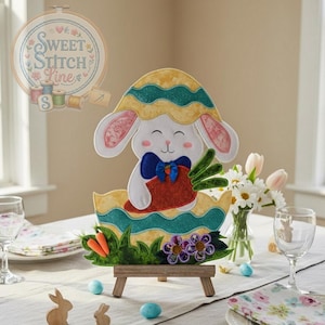 Pode incluir: Decoração de Páscoa em feltro com um coelhinho sorridente saindo de um ovo decorado. O coelhinho usa um colete vermelho e uma gravata borboleta azul, segurando cenouras. O ovo é amarelo e turquesa, colocado em um cavalete de madeira. O texto "Sweet Stitch Line" está no fundo.