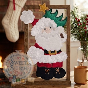 May include: A framed Christmas decoration featuring a Santa Claus design. The Santa is dressed in a red suit with white trim, a black belt, and black boots. He holds a green Christmas tree and is topped with a gold star.