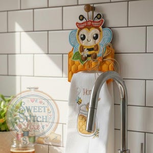 Op de afbeelding: Een decoratieve keukendoekhouder met een bijenontwerp. De houder is geel met een honingraatpatroon en houdt een witte handdoek met een honingpot en bijenborduurwerk vast. De handdoekhouder is bevestigd aan een houten peg.