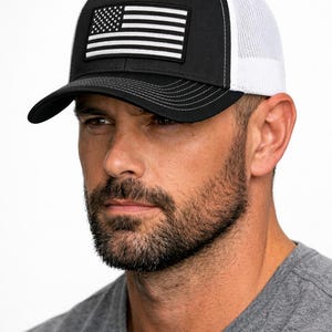 May include: A black and white trucker cap featuring a black front panel with a rectangular patch displaying a monochromatic American flag design. The cap includes a curved peak with visible stitching and a white mesh back panel for ventilation.
