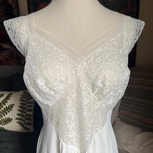 Könnte beinhalten: Ein weißes Vintage-Unterkleid auf einer Schaufensterpuppe. Das Oberteil hat eine zarte Spitze mit V-Ausschnitt und Flügelärmeln. Der Rock ist aus fließendem, einfarbigem weißem Stoff. Ein klassisches Wäschestück.