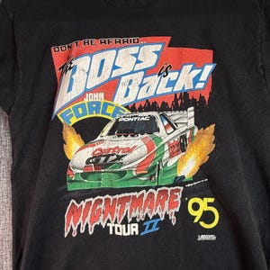 May include: A black t-shirt featuring a graphic of a Pontiac race car with flames. The text reads: DON'T BE AFRAID... THE BOSS IS BACK! JOHN FORCE CASTROL GTX NIGHTMARE TOUR II 95. The design includes red, white, green, and yellow colors.