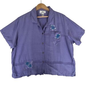 May include: A short-sleeved, button-down shirt in a light purple hue. The shirt features a collar, a front button closure, and two patch pockets. Embroidered blue floral designs are visible on the left chest pocket and the lower right pocket.
