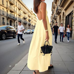 Puede incluir: Un vestido largo maxi amarillo pálido sin espalda con tirantes finos. El vestido lo lleva una persona que lleva un pequeño bolso negro. La persona camina por una calle de la ciudad con coches y peatones.
