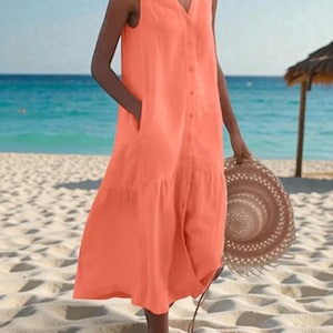 May include: A sleeveless, coral-colored dress with a V-neck and button-down front. The dress features pockets and a tiered skirt, and is worn with a straw hat. The background shows a beach with blue water and a thatched roof.