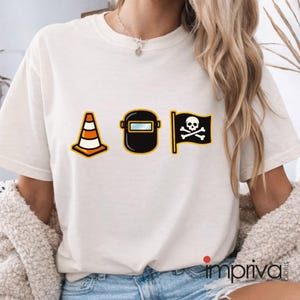 以下が含まれることがあります： クリーム色のTシャツ。オレンジと白の三角コーン、黒い溶接マスク、ドクロと骨の黒い海賊旗の3つの漫画グラフィックが描かれています。このシャツはクルーネックです。