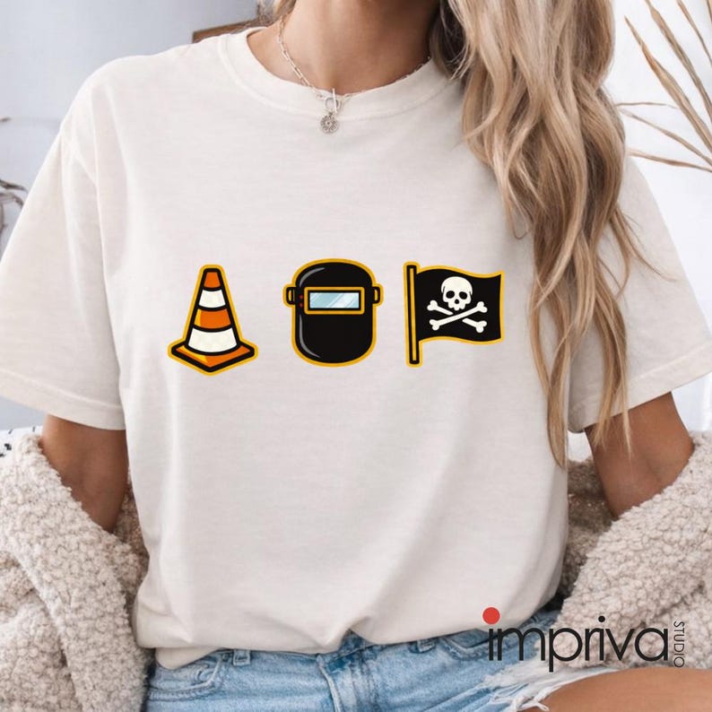 以下が含まれることがあります： クリーム色のTシャツ。オレンジと白の三角コーン、黒い溶接マスク、ドクロと骨の黒い海賊旗の3つの漫画グラフィックが描かれています。このシャツはクルーネックです。