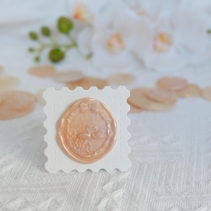 May include: A peach-colored wax seal with a floral design is centered on a white, postage stamp-shaped card. Scattered around are similar wax seals and white flowers, all on a textured white surface. The image suggests a theme of stationery or crafting.