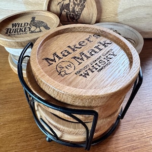 May include: A set of wooden coasters with various designs and text, including "Maker's Mark Whisky", a bison, and "Wild Turkey Bourbon". The coasters are held in a black metal holder and are made of light brown wood.