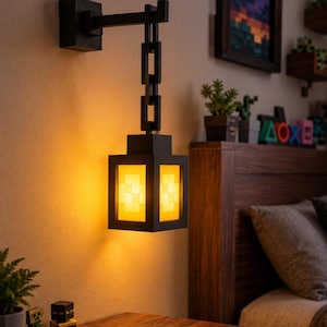Könnte beinhalten: Eine Wandlaterne mit schwarzem Rahmen, einem kettenartigen Arm und einer leuchtend gelben Lichtquelle mit pixeligem Gittermuster. Die Laterne strahlt ein warmes, sanftes Licht an die Wand und zeigt ein blockartiges, geometrisches Design.