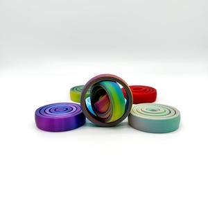 Puede incluir: Una colección de objetos circulares coloridos. Uno es un anillo con capas de colores del arcoíris dentro de un marco gris oscuro. Otros objetos son círculos planos y sólidos en tonos de morado, verde, rojo y verde azulado. Los objetos están sobre un fondo blanco.