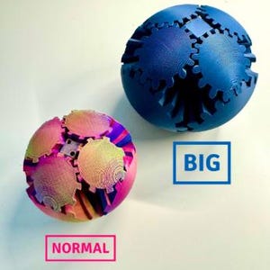 Könnte beinhalten: Zwei kugelförmige Puzzles mit Zahnrad-Design. Eines ist rosa, gelb und lila, mit der Aufschrift "NORMAL". Das andere ist eine größere, blaue Kugel, mit der Aufschrift "BIG". Beide liegen auf einer weißen Oberfläche.