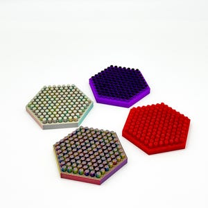 Puede incluir: Cuatro posavasos hexagonales en tonos rojo, morado, iridiscente y verde. Cada posavasos tiene una superficie texturizada con pequeñas protuberancias redondeadas. Los posavasos están dispuestos sobre un fondo blanco.