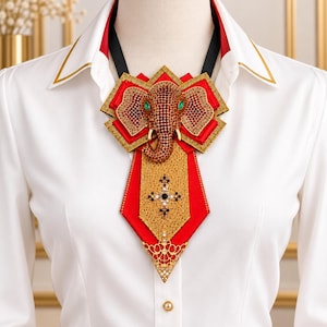 May include: A white collared shirt featuring a decorative neck accessory with a central elephant motif. The piece is crafted from red and gold fabric, adorned with brown crystals, green accents, and a cross-shaped embellishment with pearls.