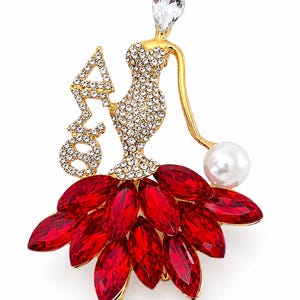 May include: A gold-toned brooch in the shape of a woman wearing a sparkling dress and a skirt of red gems. The brooch features a large clear crystal at the top and a pearl. The letters "ΑΣΦ" are also included.