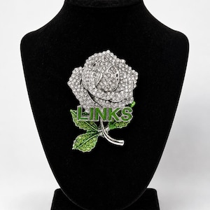 May include: A silver-toned rose brooch encrusted with clear rhinestones, featuring green rhinestone-covered leaves and the word LINKS in silver capital letters across the center of the flower. The brooch is displayed on a black velvet bust.
