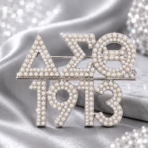 May include: A silver-toned brooch featuring the Greek letters Delta, Sigma, and Theta, followed by the number 1913. The entire surface of the letters and numbers is encrusted with small, white, round faux pearls, creating a textured, beaded appearance.