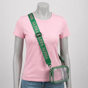 May include: A clear rectangular crossbody bag with green trim and a matching green adjustable strap. The strap features the text "1908 ALPHA KAPPA ALPHA" in light pink lettering. The bag has silver-toned metal hardware and a zipper closure.