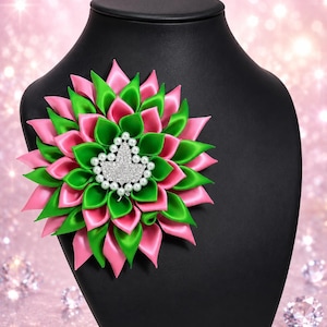May include: A decorative floral brooch in pink and green satin ribbon, adorned with a central cluster of faux pearls and rhinestones. The brooch is displayed on a black mannequin, set against a sparkling pink background.