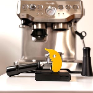 Puede incluir: Máquina de espresso de acero inoxidable con un portafiltro negro, un accesorio amarillo en forma de grano de café y una báscula negra. La máquina tiene múltiples botones y un manómetro.